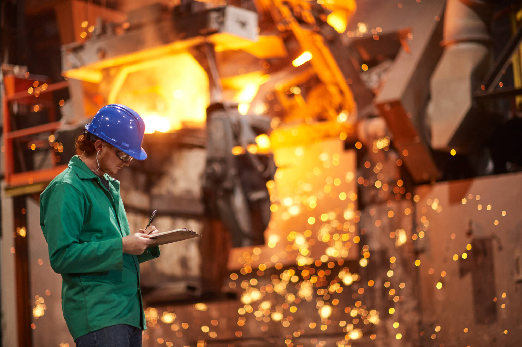A worker in a blue hard hat and green jacket records data while standing near glowing molten metal and flying sparks in a foundry.